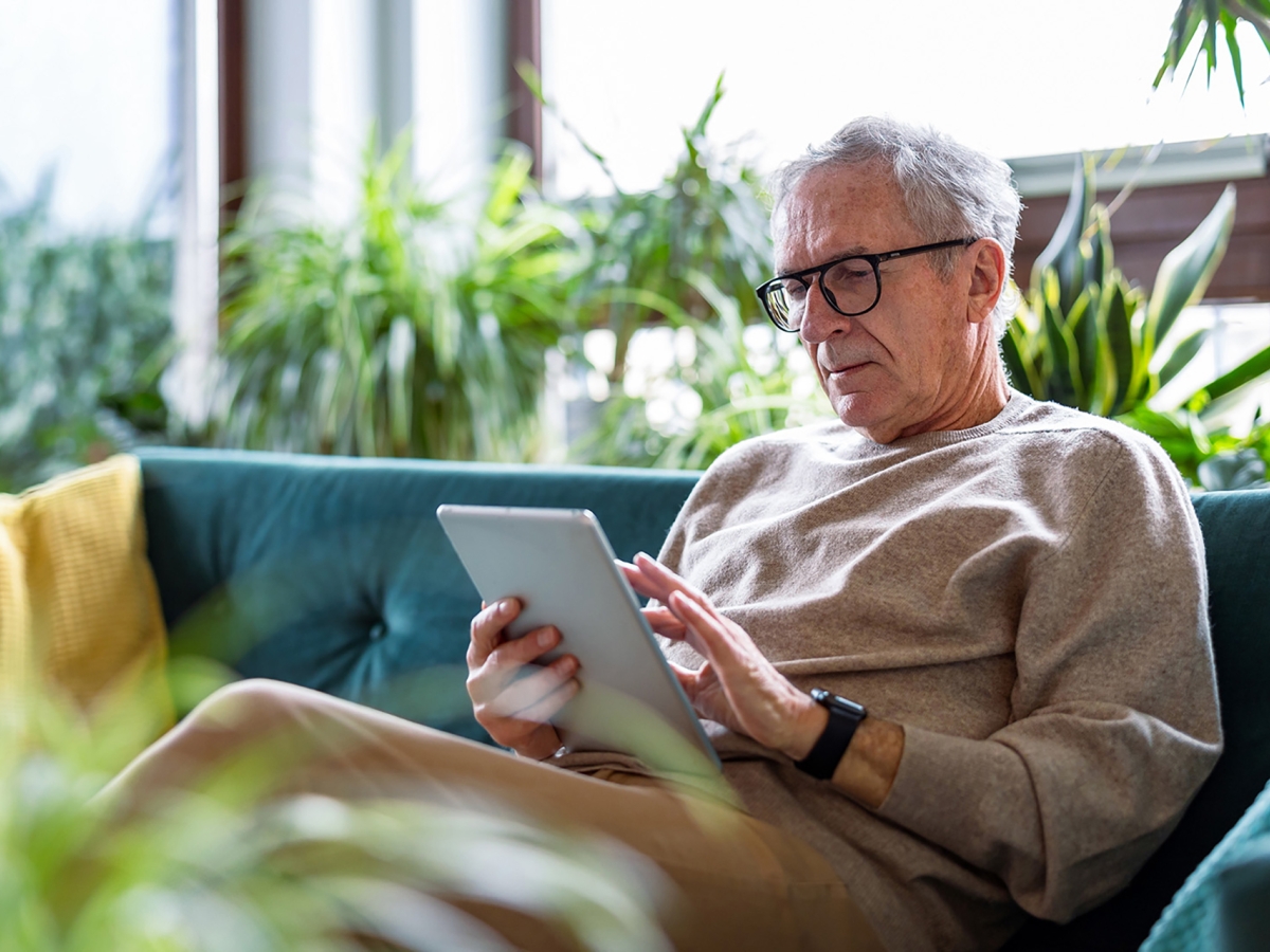 An older man sitting on a sofa, looking at his tablet in a relaxed posture.