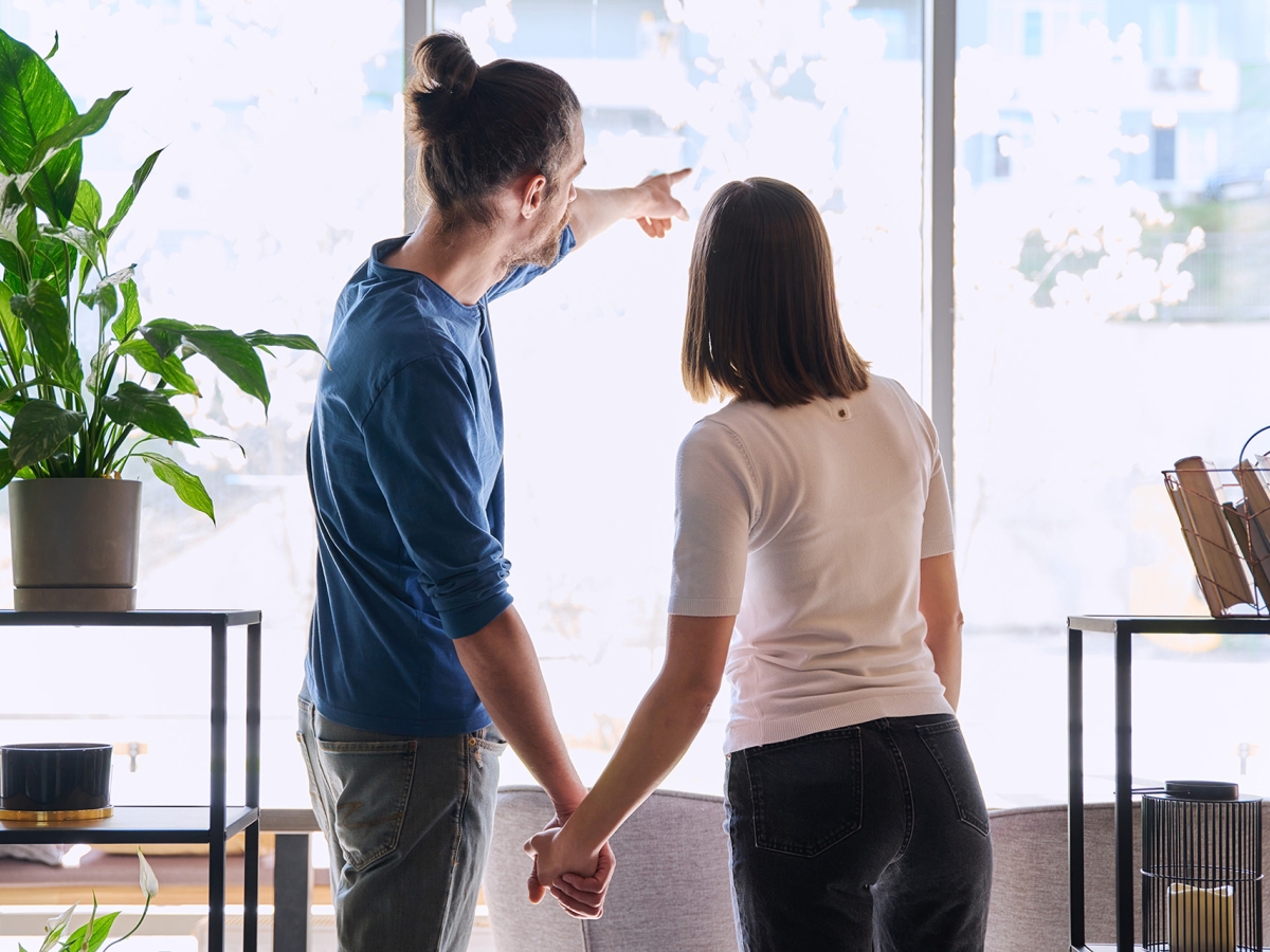 A young couple standing together, facing a window and looking out. The man is pointing towards something outside.